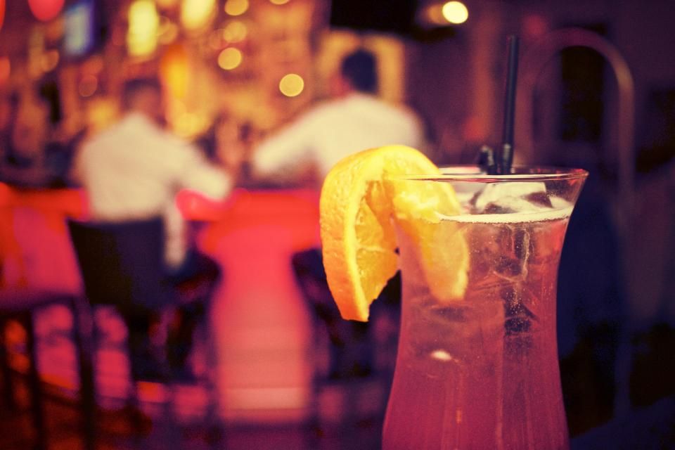 Longdrink mit Orangenscheibe im Vordergrund, dahinter Gäste an einer beleuchteten Bar