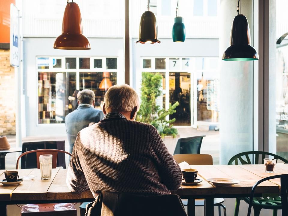 Älterer Mann sitzt mit einer Tasse Kaffee an einem Holztisch am Fenster eines Restaurants