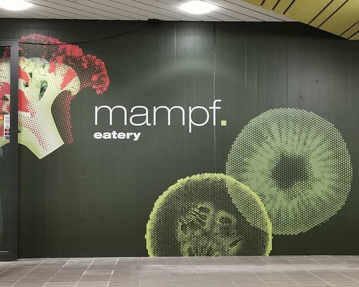 Eingang der Kantine Mampf mit dem Schriftzug «mampf. eatery» auf einer dunkelgrünen Wand mit Gemüsemotiven