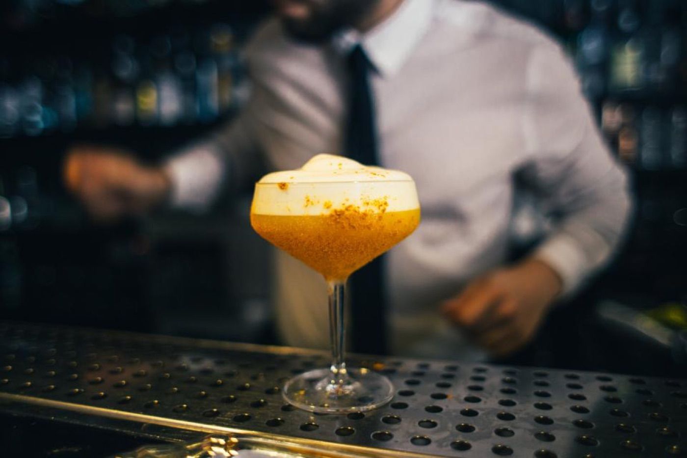 Cocktail mit Schaumkrone auf dem Bartresen, dahinter der Barkeeper