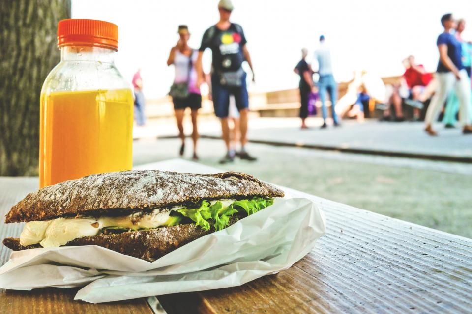 Sandwich im Papier und eine Flasche Orangensaft auf einer Holzbank, im Hintergrund Passanten